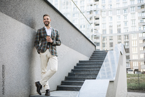 Full length portrait of joyful businessman enjoying hot beverage while relaxing outdoor. He is standing on steps and laughing 