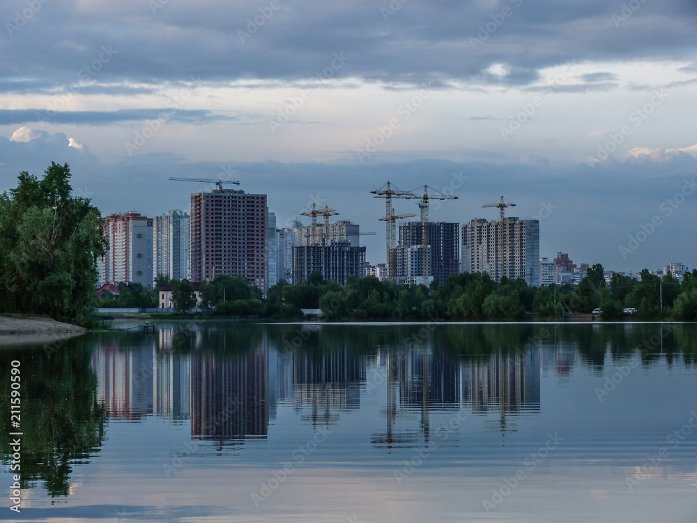 new buildings over the lake, the last urban greenery and reflections in the water, unfinished high-rise buildings for monolithic concrete technology, high-rise building cranes, poplar fluff in water