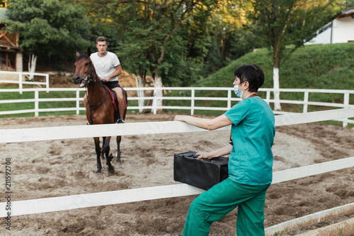 Veterinarian woman standing in front of horse coral and watching at injured and recovering animal with horseman