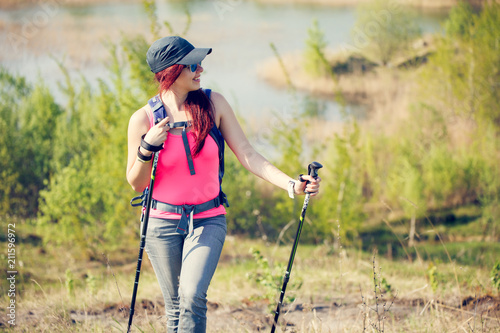 Image of young sporty girl looking away with walking sticks on background of lake and green vegetation