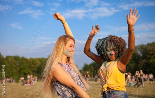 Two multiethnic girls covered in colorful powder dancing and celebrating summer holi festival