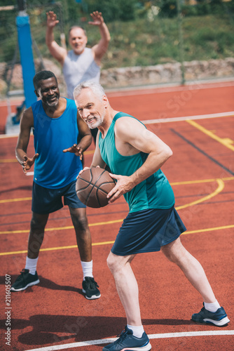 selective focus of old man with basketball ball in hands and multiracial friends behind on playground