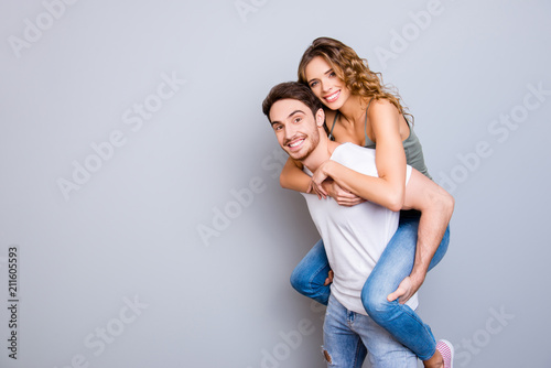 Portrait with copysapce of attractive lovely couple, strong man carrying on back charming woman with curly hair looking at camera isolated on grey background