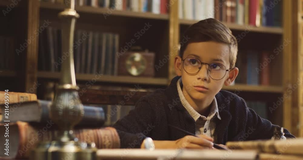 Close up of the cute teen boy in glasses writing a letter with an ink while sitting at the table in the ancient library.