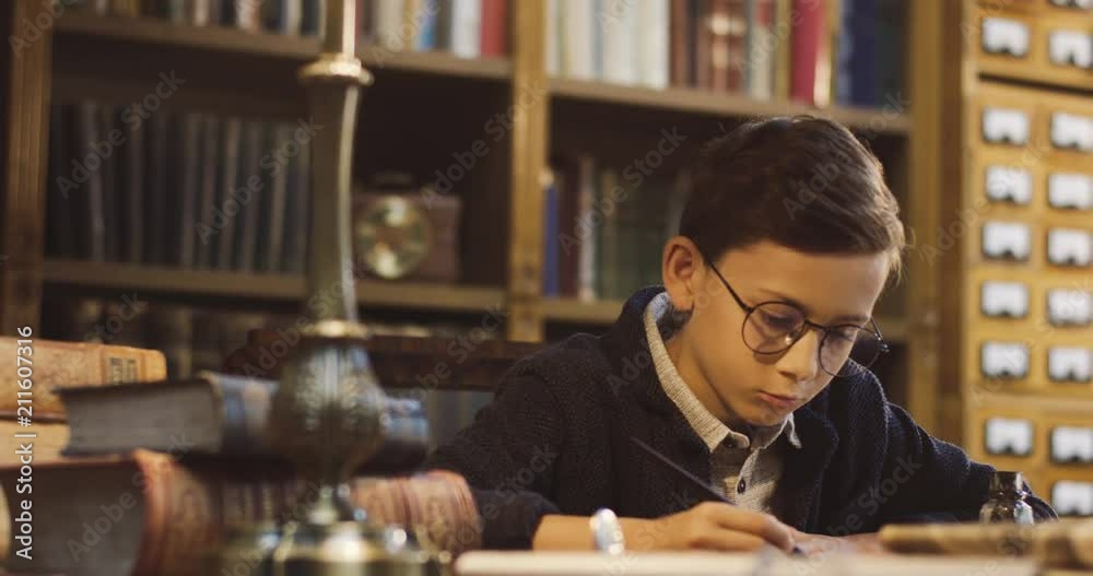 Portrait of the nice teen schoolboy in glasses writing something with an ink while sitting at the desk in the ancient library room. Calligraphy. Portrait of the nice teen schoolboy in glasses writing something with an ink while sitting at the desk in the ancient library room. Calligraphy.