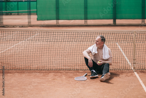 elderly man with towel and tennis equipment resting near net on tennis court