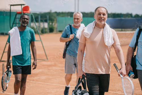 selective focus of multiracial elderly friends with tennis equipment walking on court