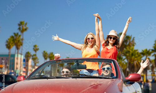 leisure, road trip, travel and people concept - happy friends driving in convertible car at country and waving hands over venice beach background in california
