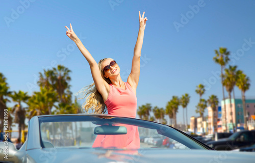travel  summer holidays  road trip and people concept - happy young woman wearing sunglasses in convertible car showing peace hand sign over venice beach background in california
