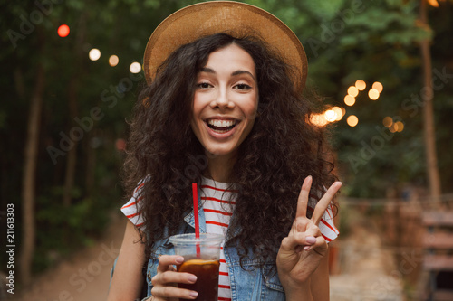 Portrait closeup of brunette beautiful woman 18-20 wearing straw hat  drinking cold tea from plastic cup and showing peace sign during rest in green park