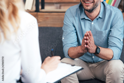 cropped image of female counselor writing in clipboard and smiling male holding hand palms together during therapy session in office photo
