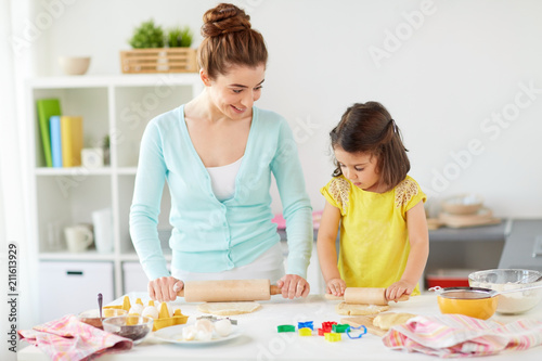 family, cooking and people concept - happy mother and little daughter with rolling pins making cookies from dough at home kitchen
