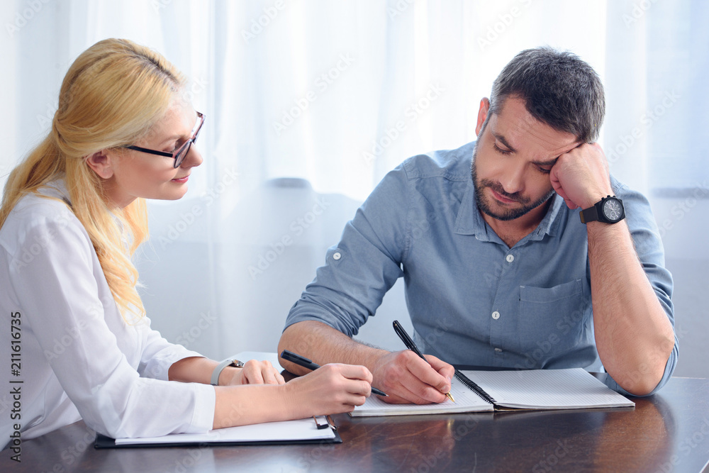 man writing down in empty textbook while sitting at table near female counselor in office man writing down in empty textbook while sitting at table near female counselor in office