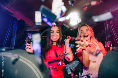 Beautiful people are sitting in car and parting. They are throwing confetti on camera. Young women are holding glasses of champagne. They are very happy and excited. photo