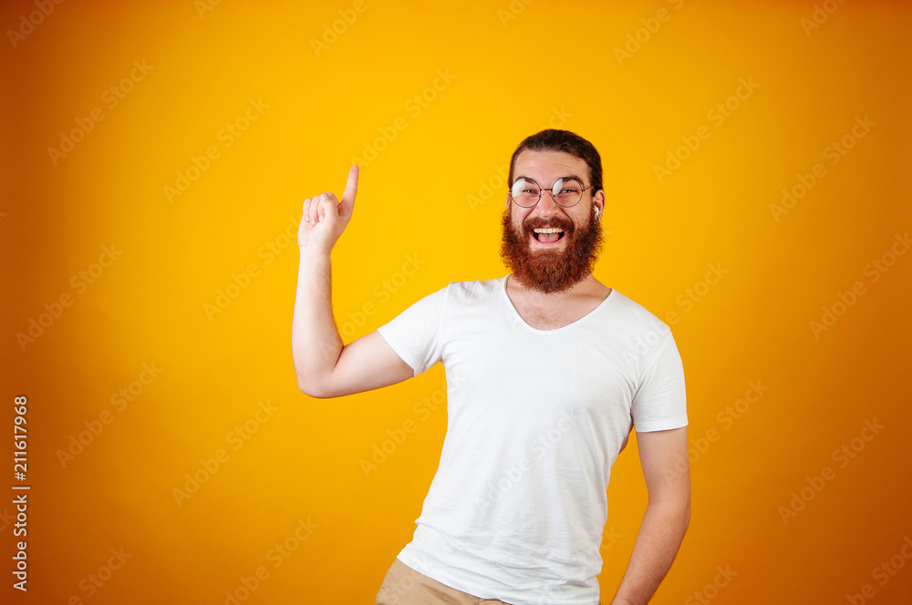 Cheerful young bearded man poting up to text space. Man wearing round framed glasses and a white T-shirt on a yellow orange background. Optics or offer concept. Cheerful young bearded man poting up to text space. Man wearing round framed glasses and a white T-shirt on a yellow orange background. Optics or offer concept.
