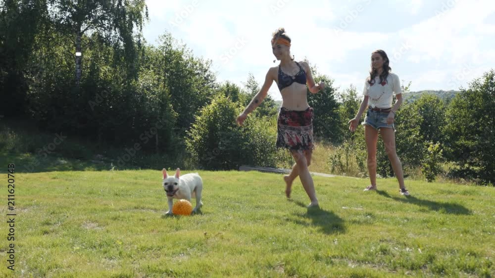 Pretty young woman playing with her pet, a friend of her in a boho style swimsuit comes outside, the dog runs for a ball straight to camera. Relaxing, having a walk. Cheerful mood. Pretty young woman playing with her pet, a friend of her in a boho style swimsuit comes outside, the dog runs for a ball straight to camera. Relaxing, having a walk. Cheerful mood.