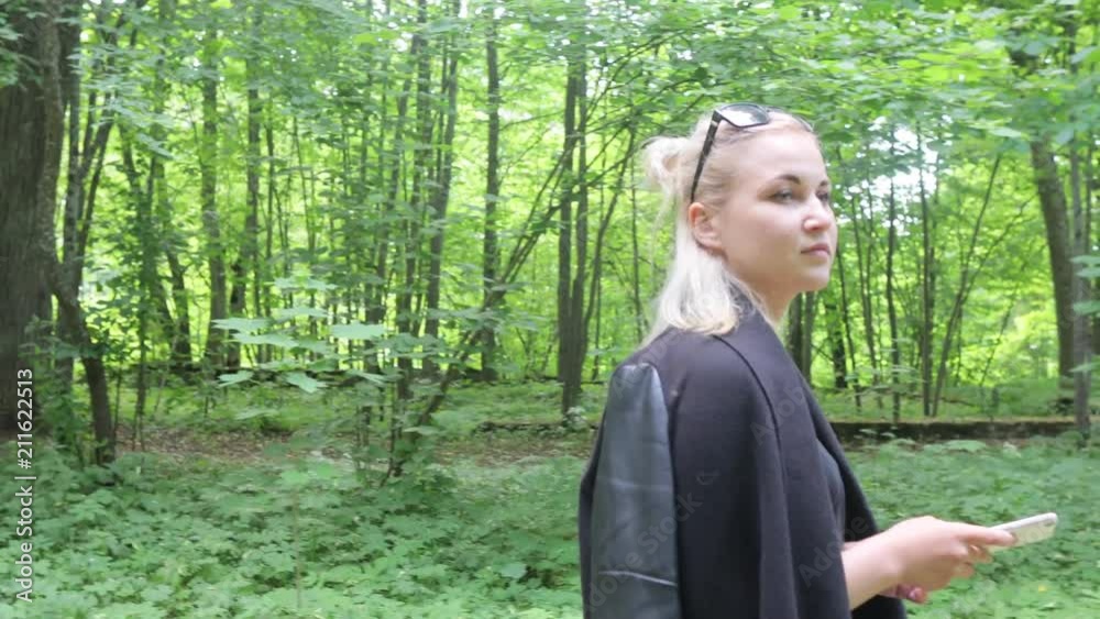 A girl with a mobile in her hands posing on the camera in the park on the road. A further shot. She is dressed in a blue skirt and black robes. A girl with a mobile in her hands posing on the camera in the park on the road. A further shot. She is dressed in a blue skirt and black robes.