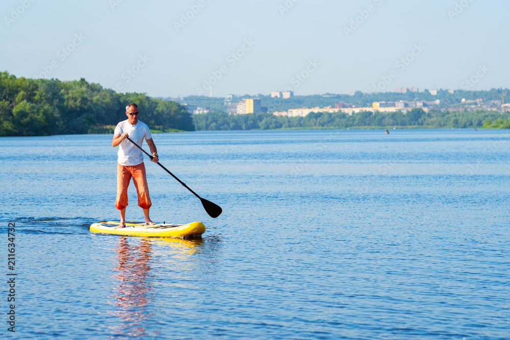 Naklejka premium Man sails on a SUP board in large river on the cityscape background