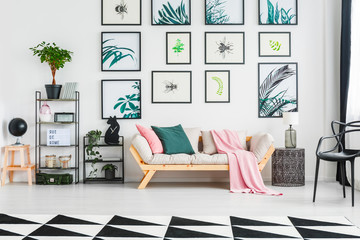 Real photo of a sofa with cushions and blanket standing between a small table with lamp and two shelves with ornaments and plants in living room interior with botanic paintings on a wall