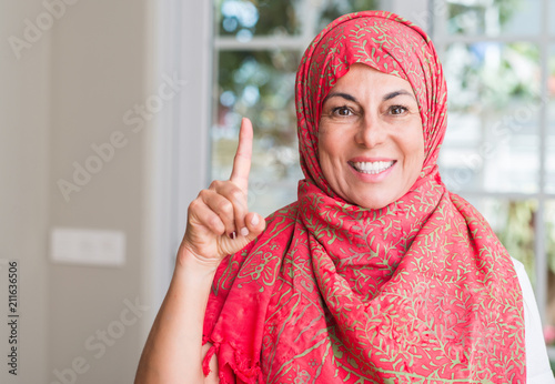 Middle aged muslim woman wearing hijab surprised with an idea or question pointing finger with happy face, number one