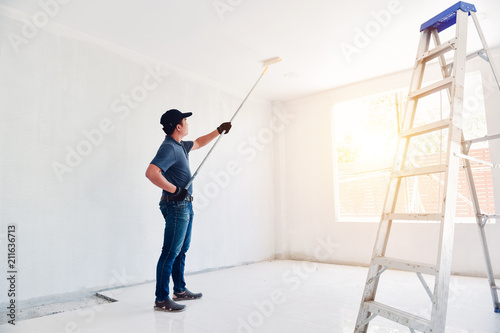 Asian rear view of a male painter drawing a wall with paint roller and a separate tank from a large empty space with wooden stairs. photo