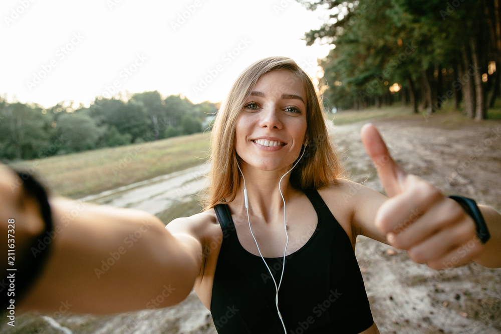 Smiling beautiful girl in sportswear taking a selfie, showing a big finger,  looking at camera. Outdoors. Stock Photo | Adobe Stock