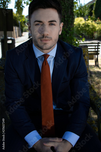 Handsome businessman with blue eyes and suit sits on village green bench at lunch time