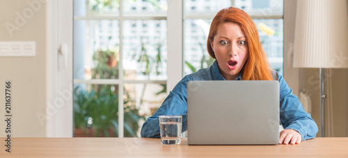 Redhead woman using computer laptop at home scared in shock with a surprise face, afraid and excited with fear expression photo