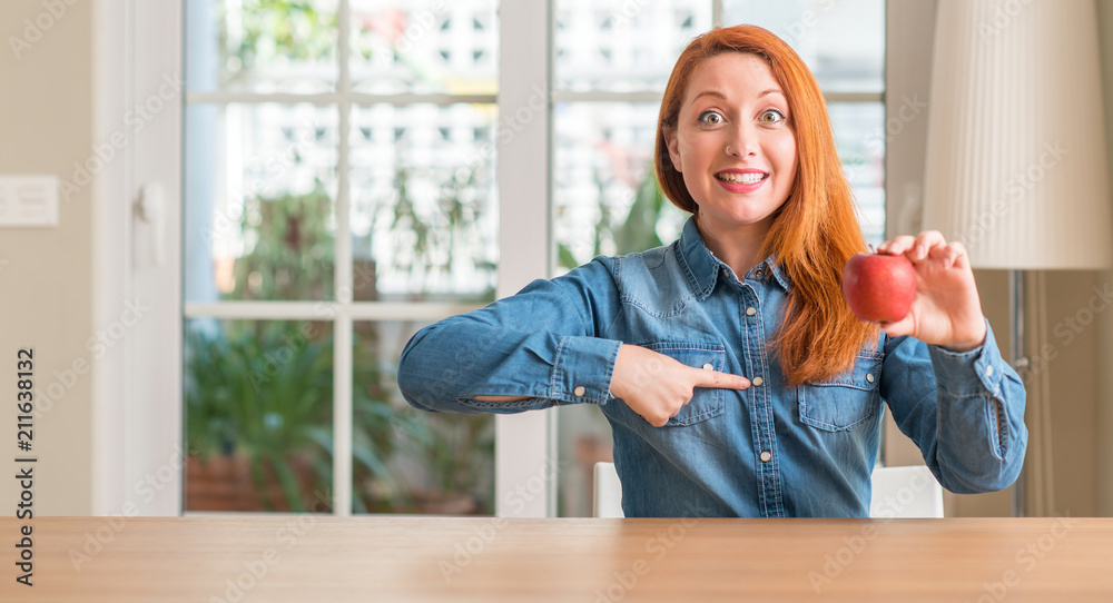 Redhead woman holding red apple at home with surprise face pointing finger to himself