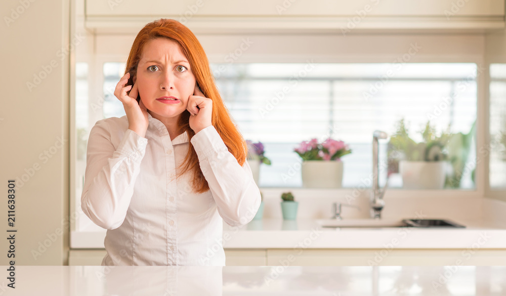 Redhead woman at kitchen covering ears with fingers with annoyed expression for the noise of loud music. Deaf concept. Redhead woman at kitchen covering ears with fingers with annoyed expression for the noise of loud music. Deaf concept.