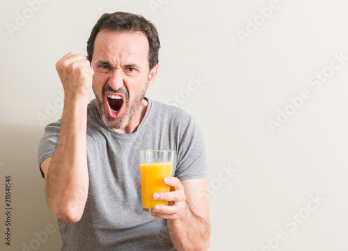 Senior man drinking orange juice in a glass annoyed and frustrated shouting with anger, crazy and yelling with raised hand, anger concept