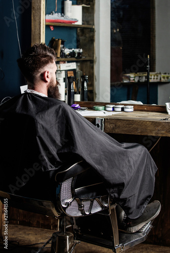 Man with beard client of hipster barbershop. Hipster with beard waits for barber and haircut. Barbershop concept. Man with beard covered with black cape sits in hairdressers chair in front of mirror