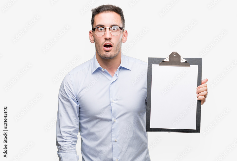 Handsome young business man showing a clipboard scared in shock with a surprise face, afraid and excited with fear expression Handsome young business man showing a clipboard scared in shock with a surprise face, afraid and excited with fear expression