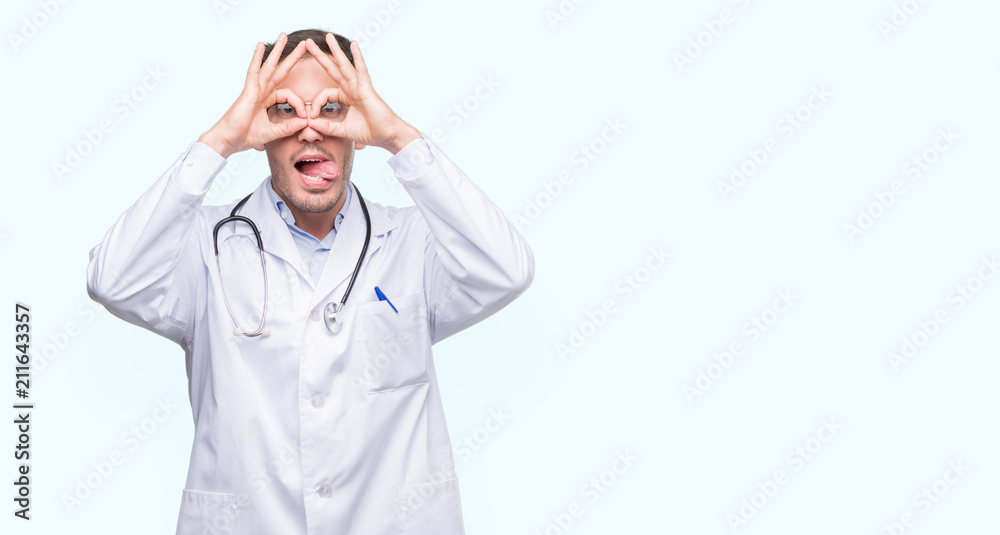 Handsome young doctor man doing ok gesture like binoculars sticking tongue out, eyes looking through fingers. Crazy expression. Handsome young doctor man doing ok gesture like binoculars sticking tongue out, eyes looking through fingers. Crazy expression.