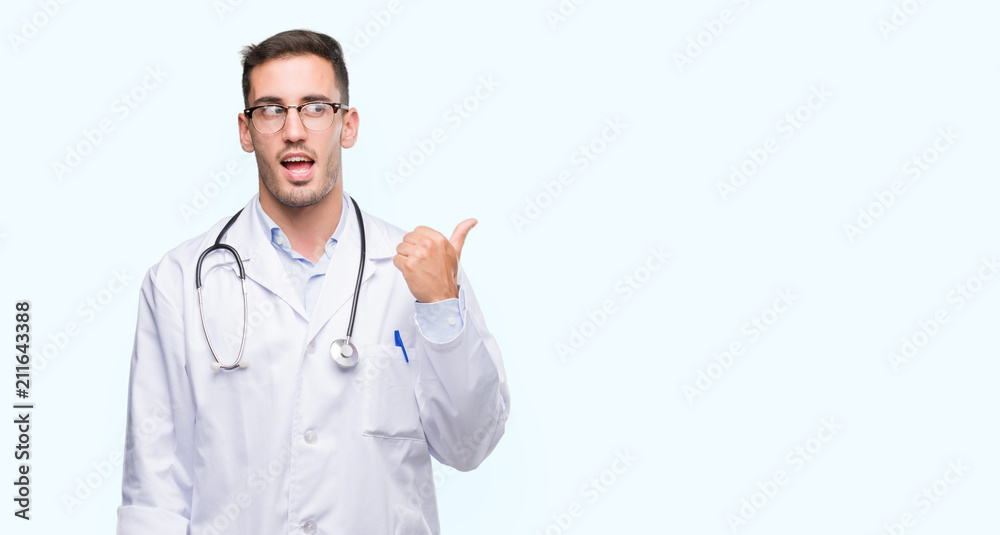 Handsome young doctor man smiling with happy face looking and pointing to the side with thumb up.