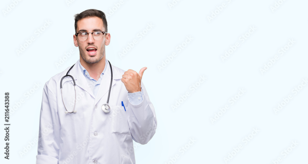 Handsome young doctor man smiling with happy face looking and pointing to the side with thumb up. Handsome young doctor man smiling with happy face looking and pointing to the side with thumb up.