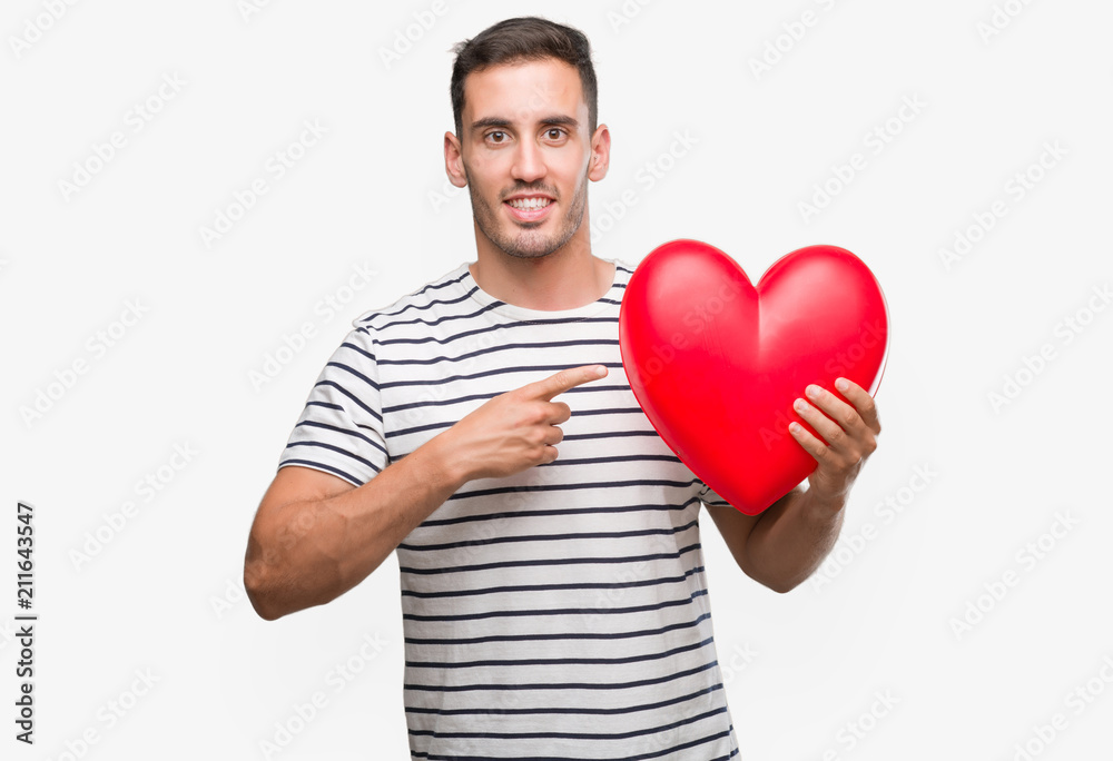 Handsome young man in love holding red heart very happy pointing with hand and finger Handsome young man in love holding red heart very happy pointing with hand and finger