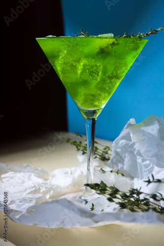 A green drink with ice cubes and green branches in a glass for mojito on crumpled wrapping paper on yellow table and blue background whth shadows photo