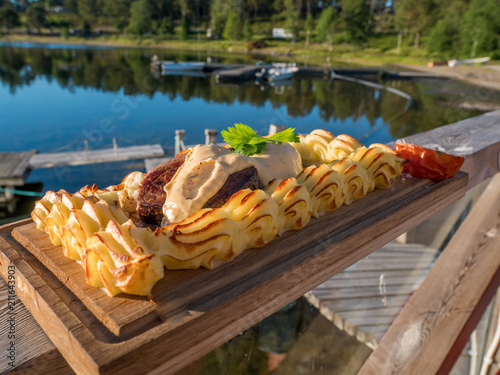 Meat steak on plank with mash potatoes puree on a wood board. Outside with lake in background. Swedish style plankstek.