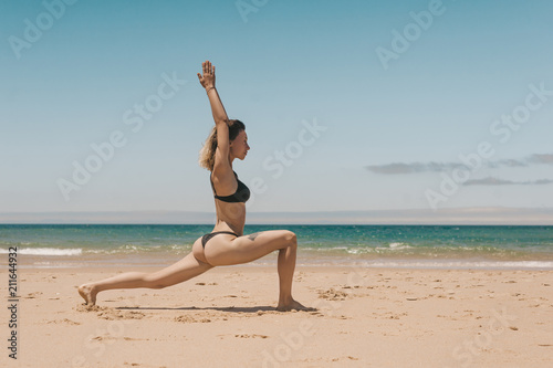 side view of young woman in black bikini standing in Warrior yoga position on sandy beach