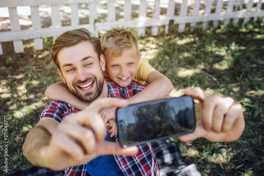 Adult and kid are looking on phone and smiling. Guy is holding device while child is hugging his dad. This picture is cute and awesome. Adult and kid are looking on phone and smiling. Guy is holding device while child is hugging his dad. This picture is cute and awesome.