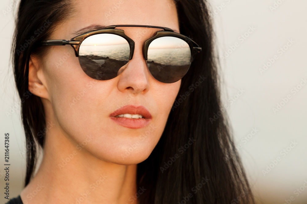 close up portrait of pretty beautiful young woman in fashionable sunglasses with reflection of beach, sunset and ocean. concept of happy holiday and resort time.