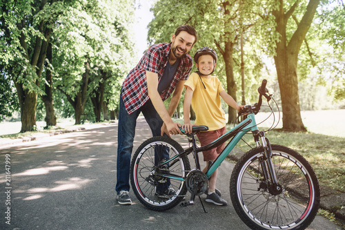 Father and son are standing and holding one bicycle. They are posing on camera and smiling. Guys are enjoying spending time together.