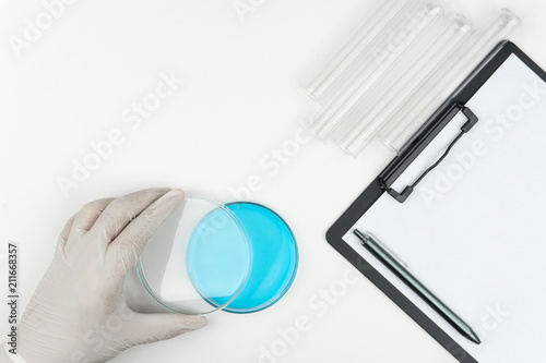 laboratory scene, the scientist opening a blue liquid watch glass, the test tubes and a clipboard with pen on the white table.