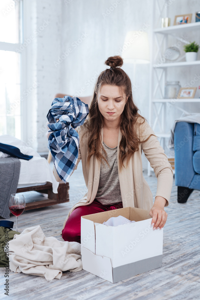 Clothes in box. Dark-haired depressed woman putting all of the clothes of ex husband into big cardboard box