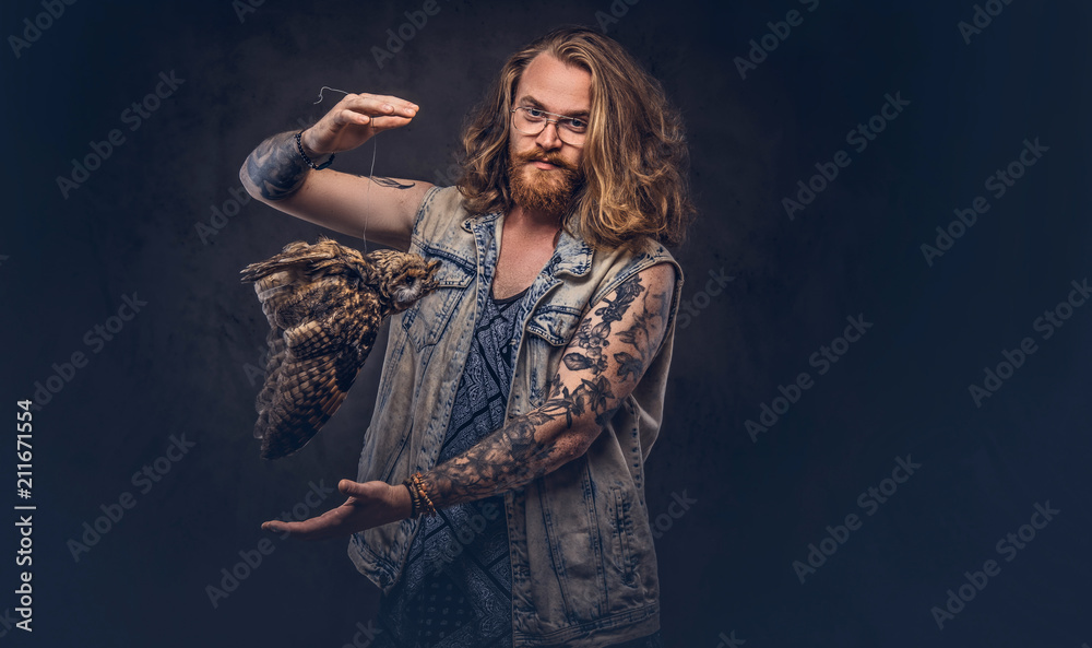 Portrait of a tattoed redhead hipster male with long luxuriant hair and full beard dressed in a t-shirt and jacket holds a keeps the scarecrow of an owl in a studio. Isolated on the dark background.