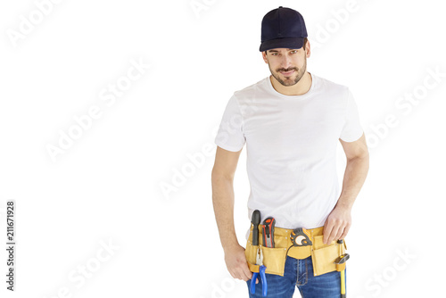 Portrait of young handyman standing at isolated white background with copy space. Successful repairman wearing baseball cap and tool belt. photo