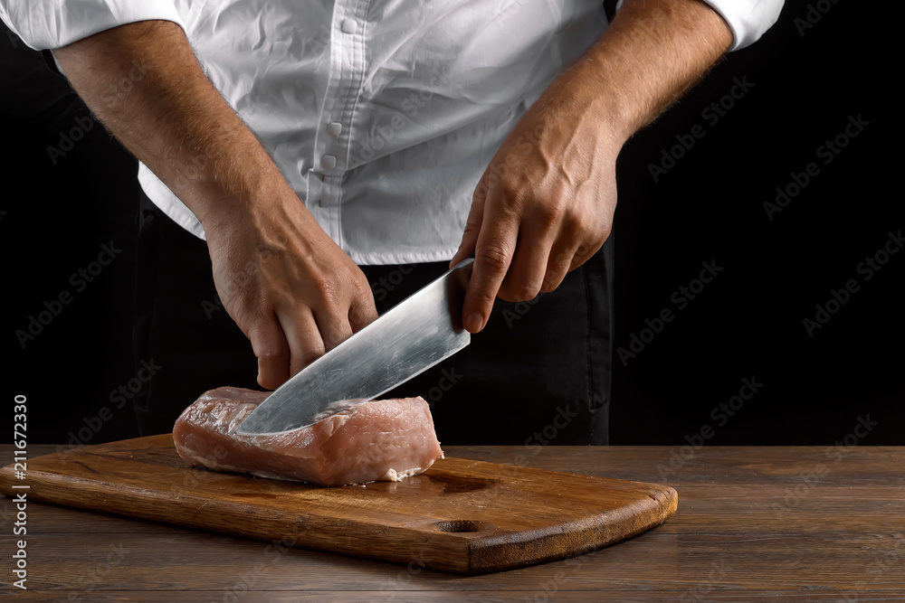 The chef cuts a piece of fresh meat on a wooden board against a dark background, hands close-up. The concept of cooking, cooking meat, a recipe for meat dishes.