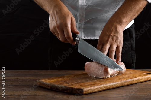 The chef cuts a piece of fresh meat on a wooden board against a dark background, hands close-up. The concept of cooking, cooking meat, a recipe for meat dishes.