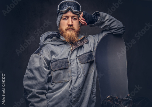 Brutal redhead snowboarder with a full beard in a winter hat and protective glasses dressed in a snowboarding coat posing with snowboard at a studio. Isolated on gray background. © Fxquadro